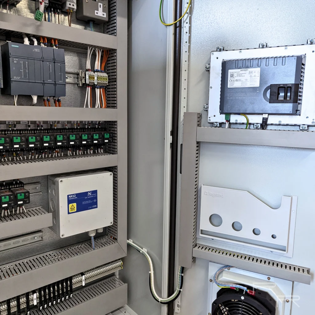 Control panel with both doors open, showing the PLC and telemetry backplate alongside the rear of a Siemens HMI, drawing pocket and ventilation fan on the door